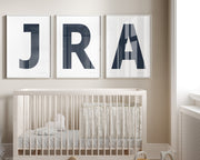 Three framed white and navy blue letter art prints spelling out a baby's initials or monogram hanging above a crib in a neutral nursery.