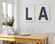 Two framed white and navy blue letter art prints spelling out LA hanging in a bright white dining room.
