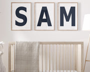 Three framed navy blue and white letter art prints spelling out the name SAM hanging above a crib in a neutral nursery.