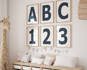 Six framed navy blue and white letter and number art prints spelling out ABC123 hanging on a wall in a nursery or playroom