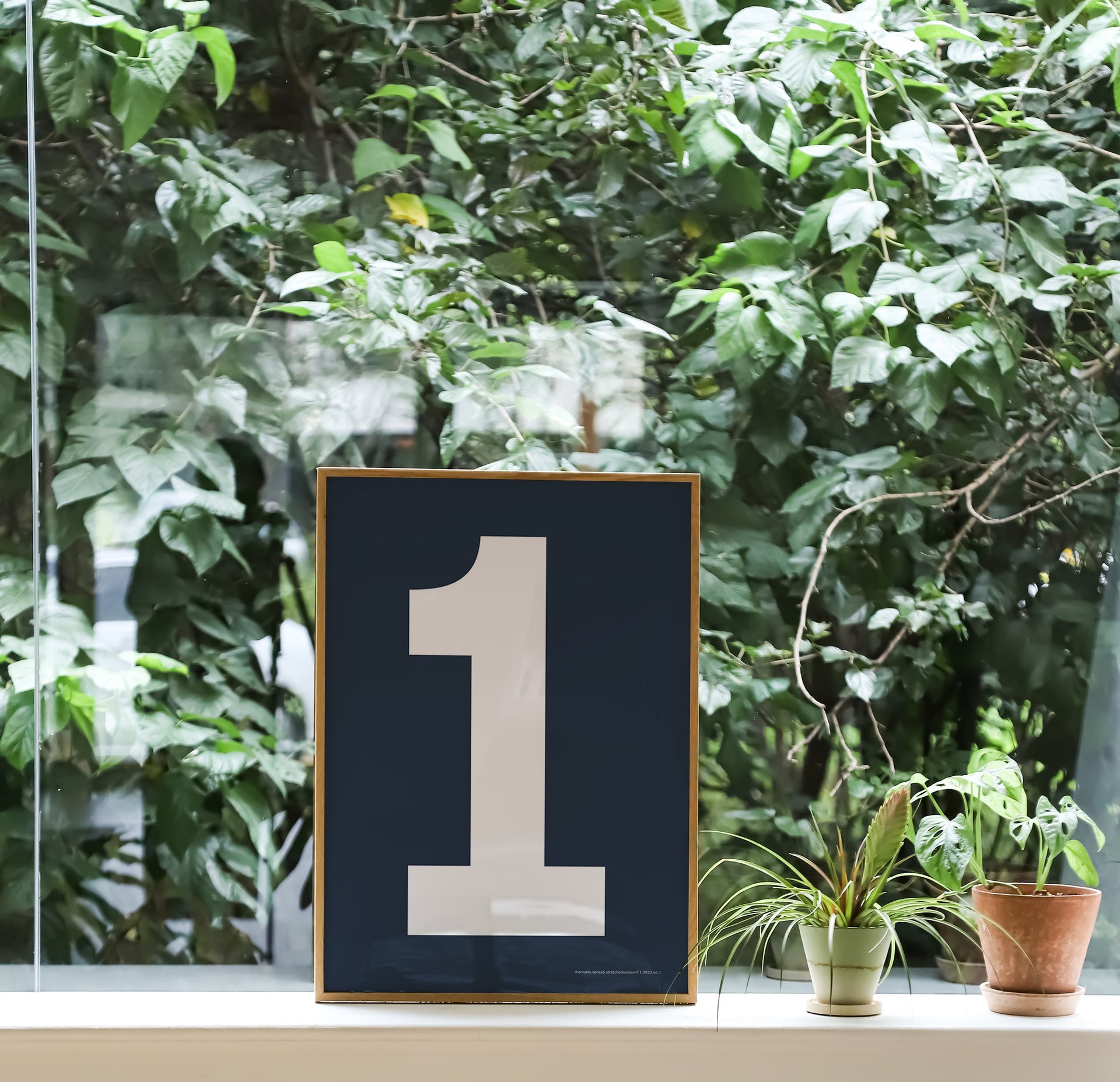 Small framed navy blue and white number 1 art print leaning against the window with green plants in the background.