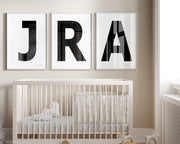 Three framed white and black letter art prints spelling out a baby's initials or monogram hanging above a crib in a neutral nursery.