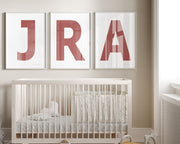 Three framed white and Nantucket red letter art prints spelling out a baby's initials or monogram hanging above a crib in a neutral nursery.