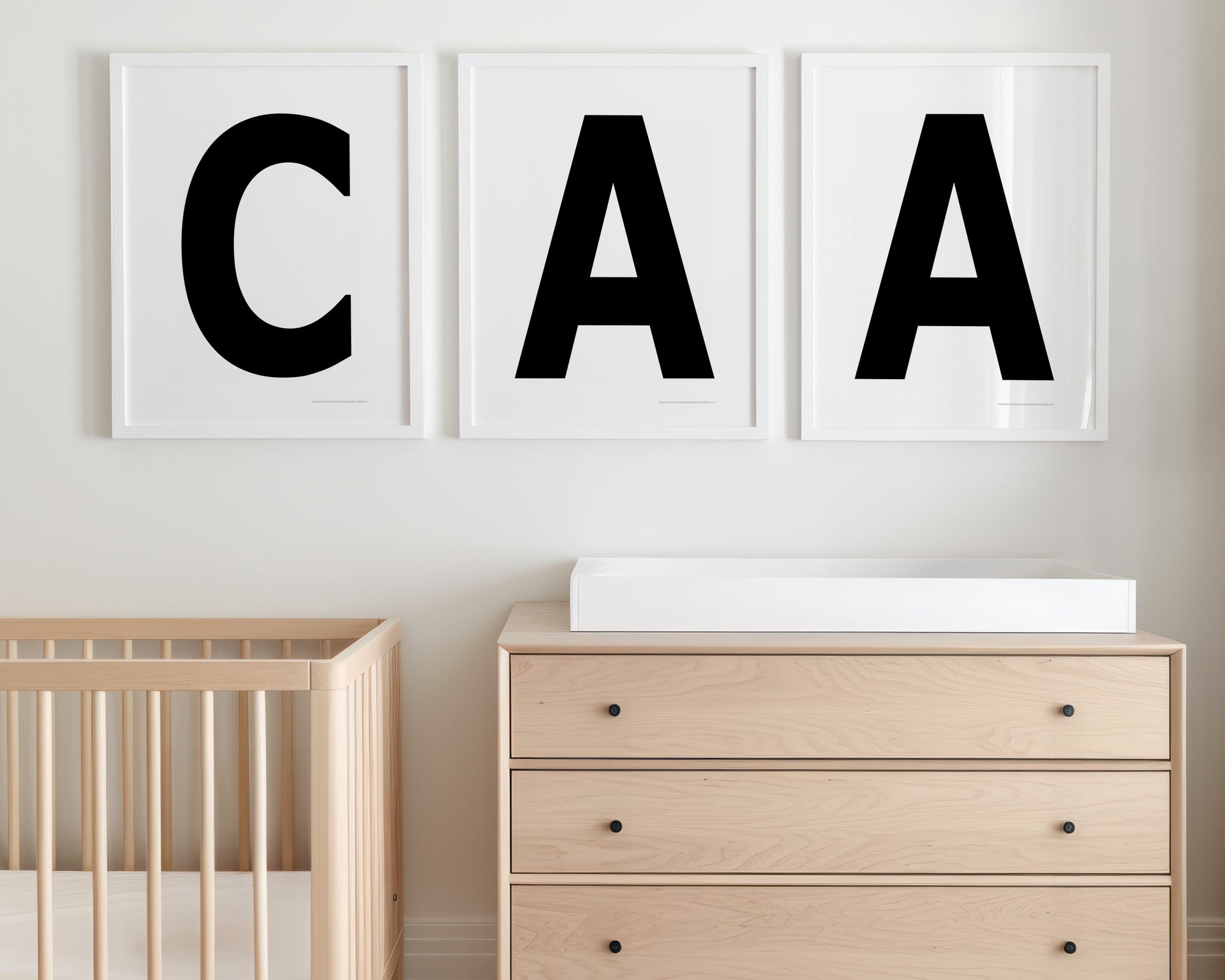 Three framed black and white letter prints featuring a baby's initials or monogram hanging above a crib and changing table in a neutral nursery.