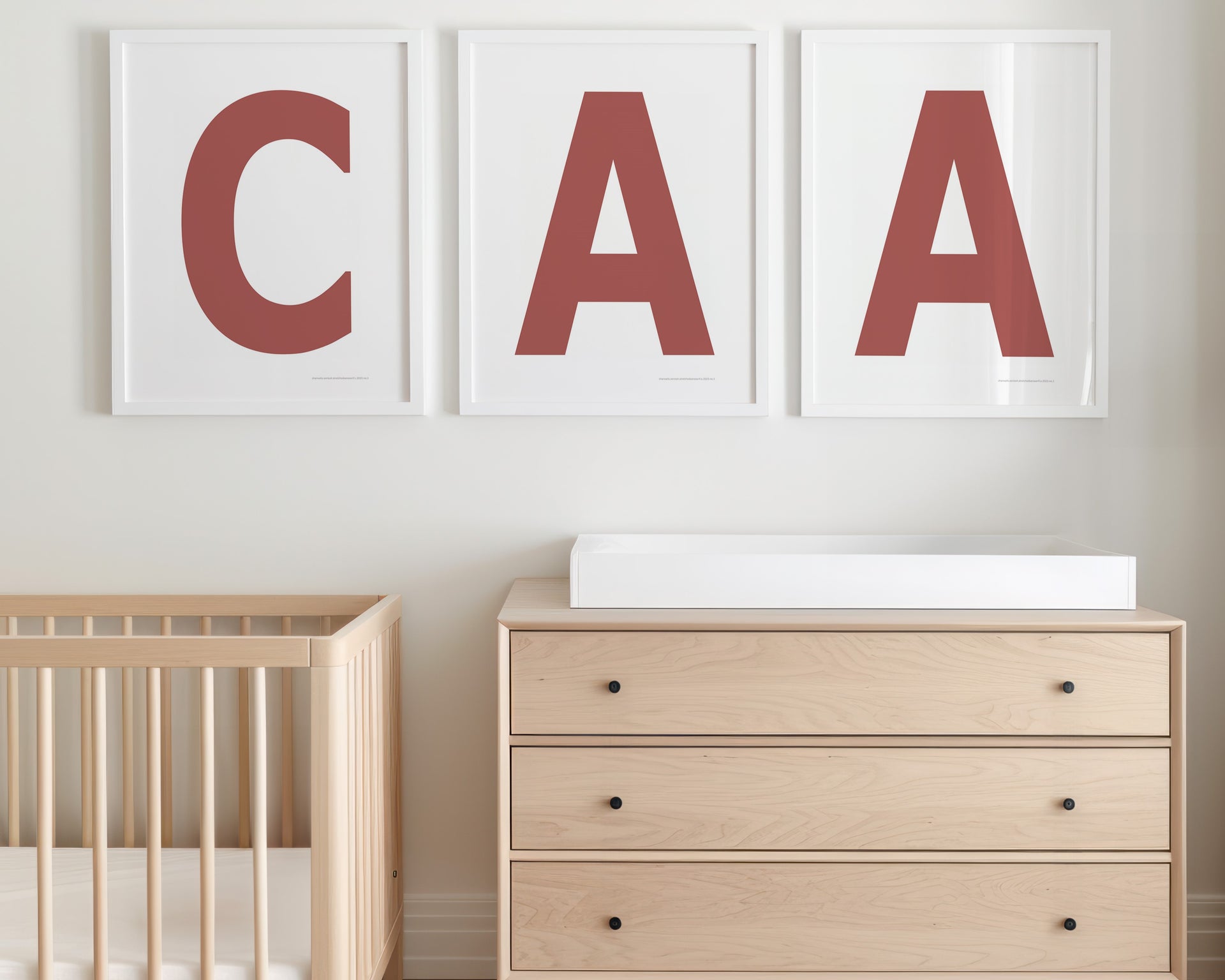 Three framed Nantucket red and white letter prints featuring a baby's initials or monogram hanging above a crib and changing table in a neutral nursery.