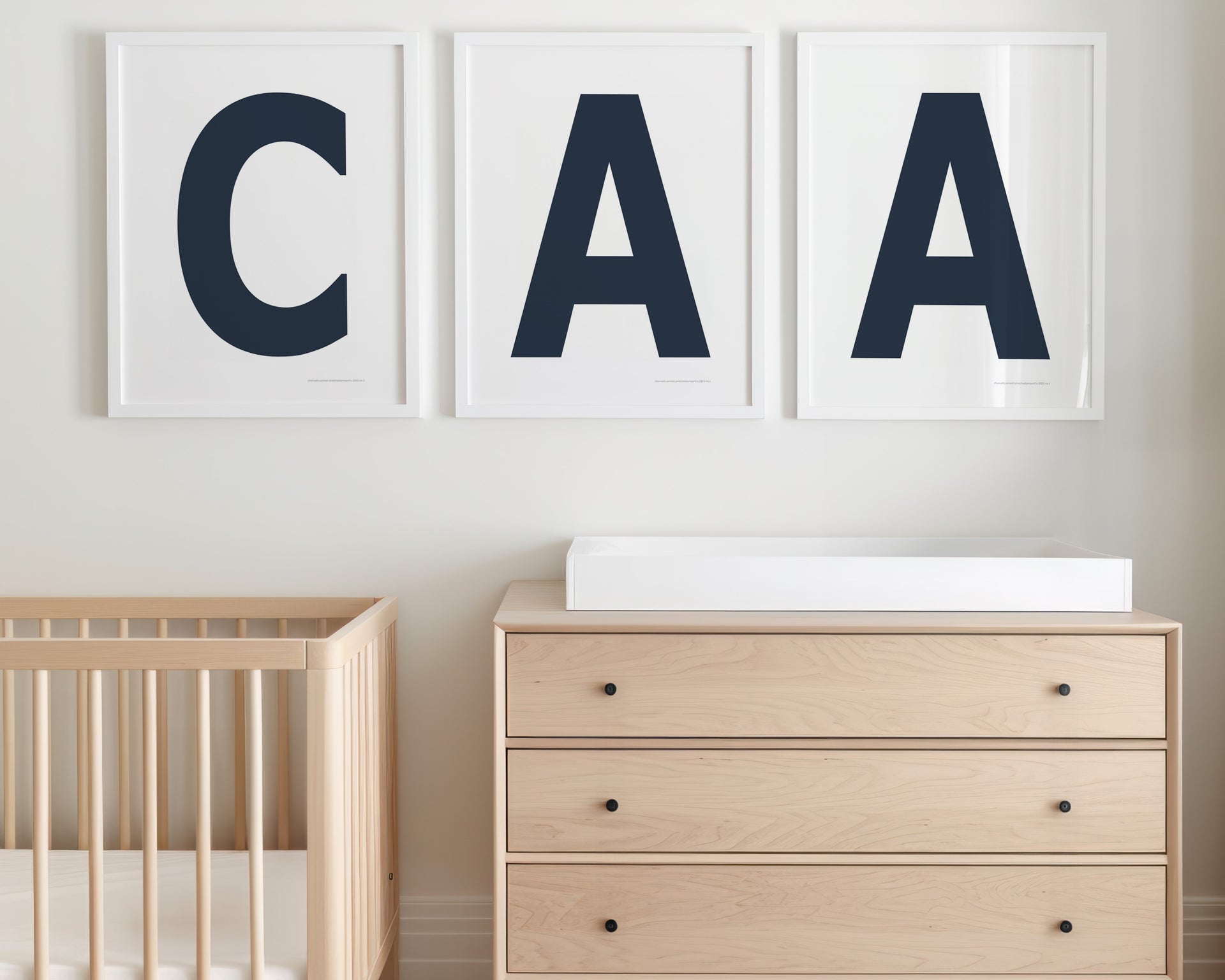 Three framed white and navy blue letter prints featuring a baby's initials or monogram hanging above a crib and changing table in a neutral nursery.