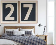 Two large framed navy blue and white number art frames showcasing a boy's jersey number hanging above his bed with a plaid blanket.