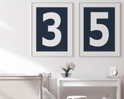 Two framed navy blue and white number art prints showing a jersey number hanging above bed and desk.