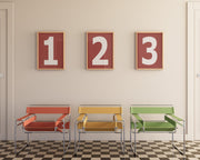 Three framed number art prints with the numbers 1, 2, and 3 hanging on wall above three modern colorful chairs.