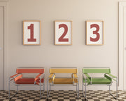 Three framed number art prints with the numbers 1, 2, and 3 hanging on wall above three modern colorful chairs.