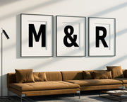 Three oversized framed black and white letter and ampersand art prints spelling out M&R hanging above a modern sofa.