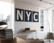 Three framed navy blue and white letter art prints spelling out NYC hanging on a wall in a modern living room with a view of the city.