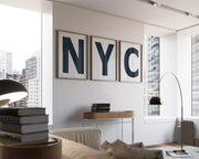 Three framed white and navy blue letter art prints spelling out NYC hanging on a wall in a modern living room with a view of the city.