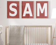 Three framed Nantucket red and white letter art prints spelling out the name SAM hanging above a crib in a neutral nursery.