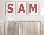 Three framed Nantucket red and white letter art prints spelling out the name SAM hanging above a crib in a neutral nursery.