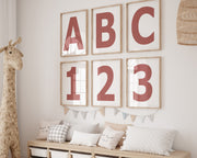 Six framed Nantucket red and white letter and number art prints spelling out ABC123 hanging on a wall in a nursery or playroom.